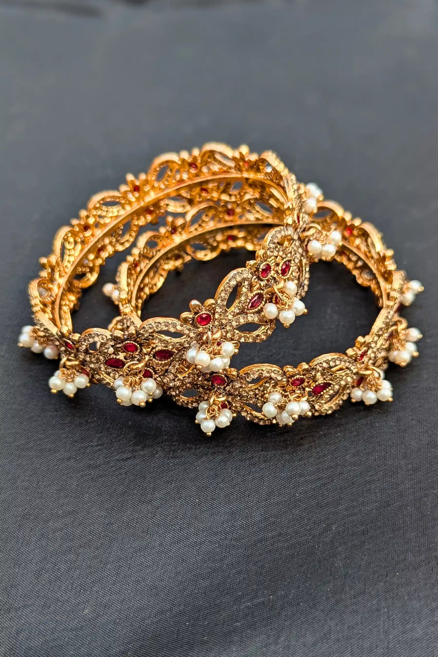 Two-piece golden bangles with red stones and pearls on a dark background