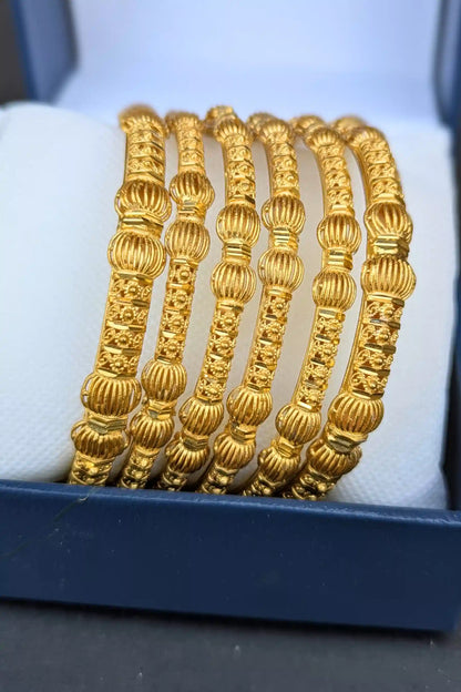 Set of gold bangles with intricate designs on a white cushion inside a blue box.