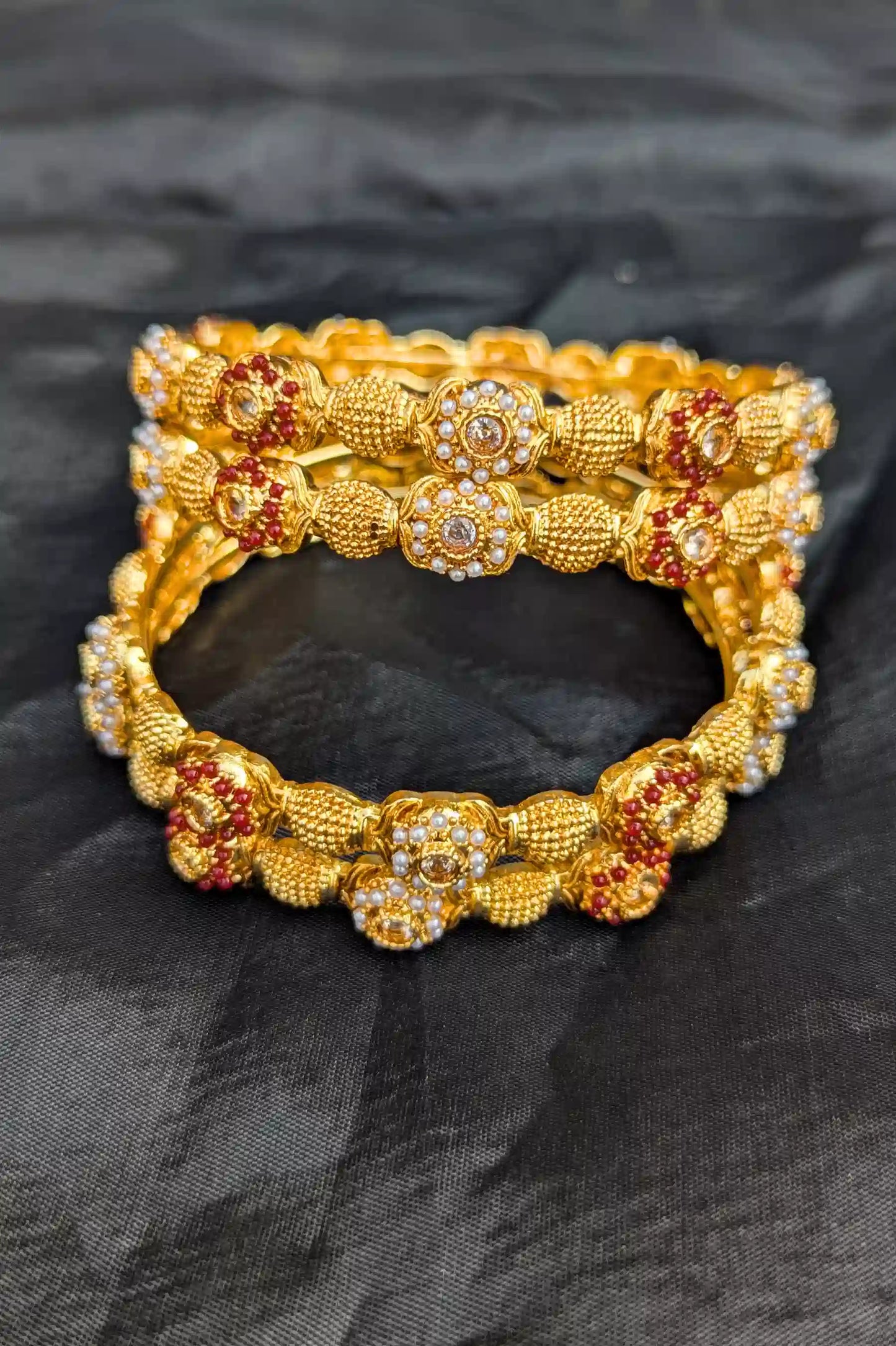 Gold bangles with floral designs and red stones on a black fabric background